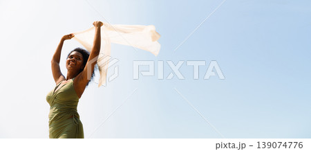 Joyful Black woman in green top stands outdoors, holding light white fabric flows in the wind against clear blue sky on the beach. Concept of freedom, mental health, and summer vacation. 139074776