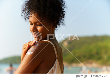 Beautiful young Black woman with curly hair smiles while applying sunscreen lotion to her shoulder on sunny beach. Concept of skin protection, summer wellness, and tropical vacation. Beautiful young Black woman with curly hair smiles while applying sunscreen lotion to her shoulder on sunny beach. Concept of skin protection, summer wellness, and tropical vacation. 139074778