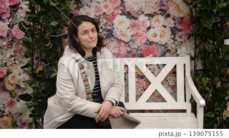 Woman sits on a white bench in front of a flower backdrop at a floral event in the afternoon Woman sits on a white bench in front of a flower backdrop at a floral event in the afternoon 139075155