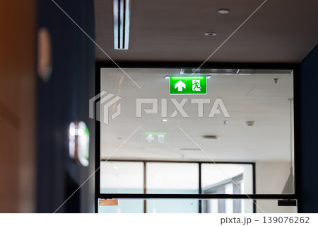 A green fire exit sign hangs from the ceiling with a running man and right arrow, combining safety direction with emergency evacuation guidance in an indoor building. A green fire exit sign hangs from the ceiling with a running man and right arrow, combining safety direction with emergency evacuation guidance in an indoor building. 139076262