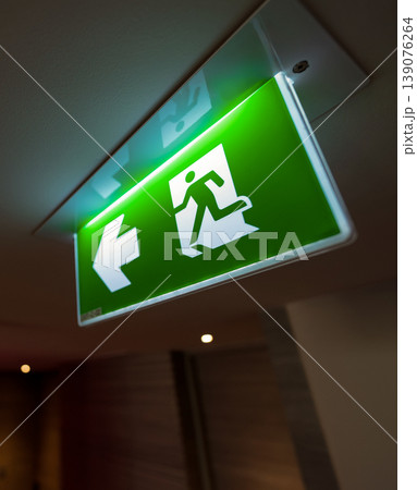 A green fire exit sign hangs from the ceiling with a running man and right arrow, combining safety direction with emergency evacuation guidance in an indoor building. A green fire exit sign hangs from the ceiling with a running man and right arrow, combining safety direction with emergency evacuation guidance in an indoor building. 139076264