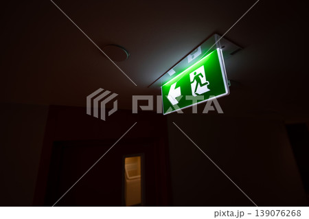 An emergency fire exit light box with arrow hangs from a hospital ceiling, combining evacuation drill concept with clear safety direction and escape guidance. An emergency fire exit light box with arrow hangs from a hospital ceiling, combining evacuation drill concept with clear safety direction and escape guidance. 139076268