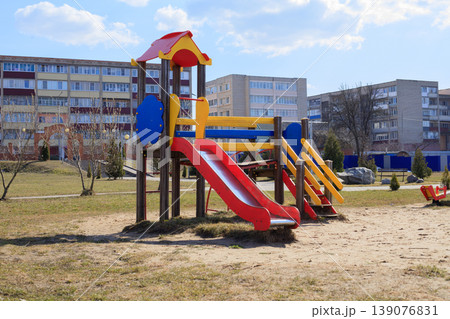 Wooden children's playground equipment with a red slide and blue climbing elements. Colorful playset in a residential area with apartment buildings Wooden children's playground equipment with a red slide and blue climbing elements. Colorful playset in a residential area with apartment buildings 139076831