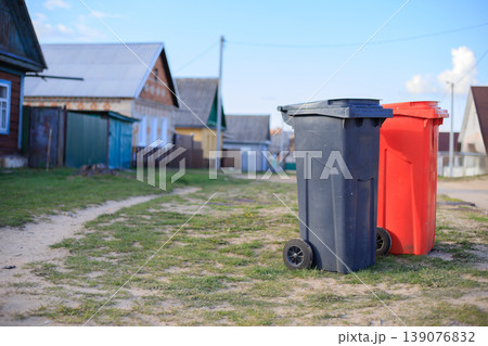 Pair of plastic garbage bins for selective waste disposal on a village street. Black and red recycling containers in a rural neighborhood. Pair of plastic garbage bins for selective waste disposal on a village street. Black and red recycling containers in a rural neighborhood. 139076832