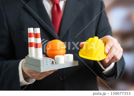 A factory and a construction helmet in the hands of a businessman. Building a new industrial plant supports modernization. Energy upgrades improve efficiency and production capacity. 139077353