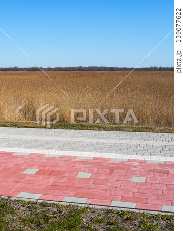 Red bicycle lane and sidewalk at edge of rural field with tall grass under clear blue sky Red bicycle lane and sidewalk at edge of rural field with tall grass under clear blue sky 139077622