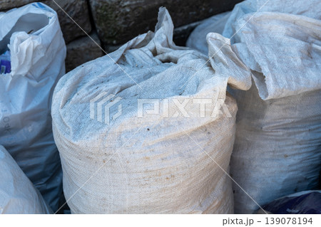 Several large bags filled with waste materials are stacked on the ground next to a stone wall in a non-urban area during the day 139078194