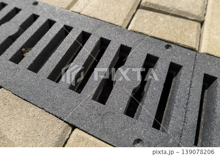 A drain cover sits on gray paving stones under bright sunlight on a clear day. The drain features several slits and holes 139078206