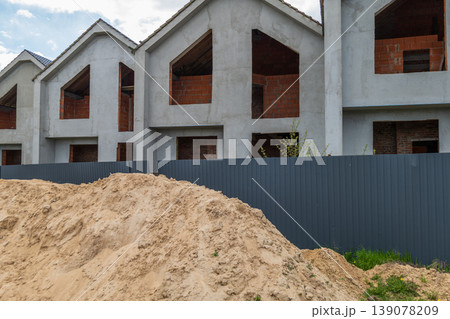 A construction site shows several houses being built with empty windows and a pile of dirt in front. The sky has clouds above the site 139078209