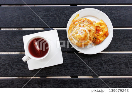 Overhead view of a cup of red fruit tea and a white plate with sweet burek pastry roll and focaccia on a dark slatted balcony table. Copy space Overhead view of a cup of red fruit tea and a white plate with sweet burek pastry roll and focaccia on a dark slatted balcony table. Copy space 139079248