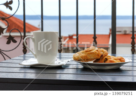 Side view of a white cup of tea and plate with burek pastry on a balcony table with the view on Adriatic sea in Makarska, Croatia on soft background. Side view of a white cup of tea and plate with burek pastry on a balcony table with the view on Adriatic sea in Makarska, Croatia on soft background. 139079251