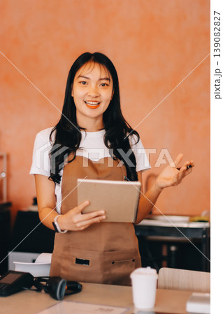 Friendly Asian female barista receives a credit card from a customer for payment. Both women are smiling in a warmly lit coffee shop with a rustic interior. Cashless payments. Small family business Friendly Asian female barista receives a credit card from a customer for payment. Both women are smiling in a warmly lit coffee shop with a rustic interior. Cashless payments. Small family business 139082827