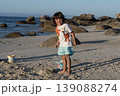 Child standing on beach with rocks and ocean background holding bucket during summer day 139088274