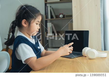 Young girl engaged with modern technology while sitting at a wooden table in a cozy learning environment with a laptop, headphones, and books nearby 139089351
