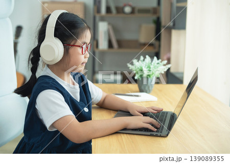 Young girl wearing headphones using laptop in bright indoor workspace with plants and books, focused on online learning or homework task 139089355