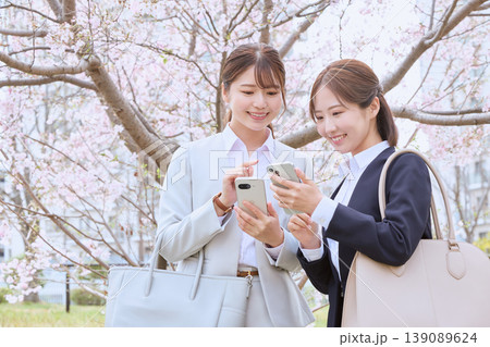 桜の下でスマートフォンを見せ合う新入社員の若い女性二人・春のビジネスシーン 桜の下でスマートフォンを見せ合う新入社員の若い女性二人・春のビジネスシーン 139089624