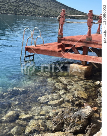 Red wooden pier over clear turquoise water with rocky shore, vertical background Red wooden pier over clear turquoise water with rocky shore, vertical background 139090052