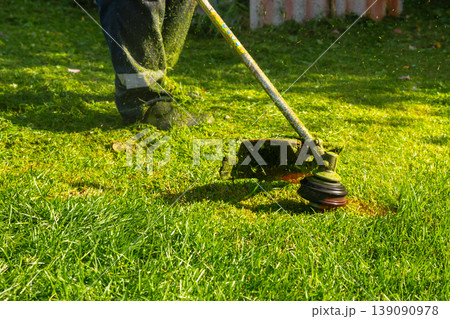 Man mowing grass with string trimmer. Gardener trimming lawn in summer. Professional landscaping and yard maintenance. Powerful tool for horticulture, backyard improvement and garden care. Man mowing grass with string trimmer. Gardener trimming lawn in summer. Professional landscaping and yard maintenance. Powerful tool for horticulture, backyard improvement and garden care. 139090978