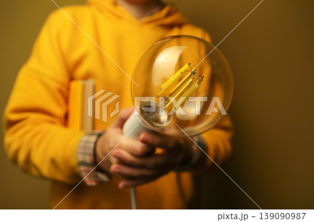 Person in yellow hoodie holding a large vintage Edison light bulb. Concept of creative idea, innovation, and inspiration. Close-up of a decorative lamp in hands with a blurred background 139090987