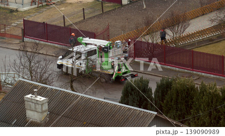 Aerial view of a utility service truck with a cherry picker platform parked on a suburban street. Workers in helmets performing maintenance near a residential fence 139090989