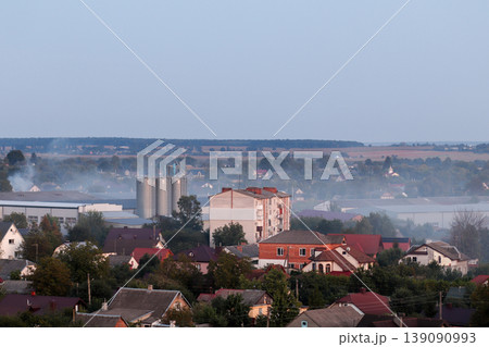 Air pollution from industrial factory pipes over a small town. Concept of environmental issues, smog, and carbon emissions affecting residential areas. Grain elevator 139090993