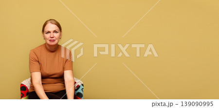 Middle-aged woman sitting in a colorful armchair against a solid yellow background with large copy space. Portrait of a calm female in a brown t-shirt. Minimalist studio photography for advertising 139090995