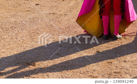 Close up of a bullfighter costume of pink stockings, black shoes, yellow and pink cape on the sand. 139091240