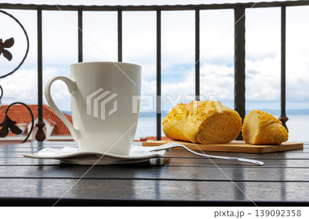 White cup of tea and croatian corn bread on a balcony table with the view of Adriatic sea in Makarska, Croatia on soft background. White cup of tea and croatian corn bread on a balcony table with the view of Adriatic sea in Makarska, Croatia on soft background. 139092358