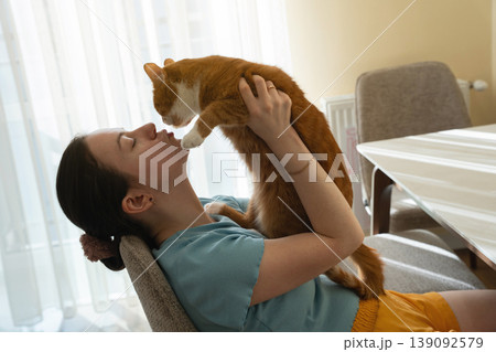 A brunette caucasian woman kisses and plays with a ginger domestic cat while sitting in a kitchen A brunette caucasian woman kisses and plays with a ginger domestic cat while sitting in a kitchen 139092579