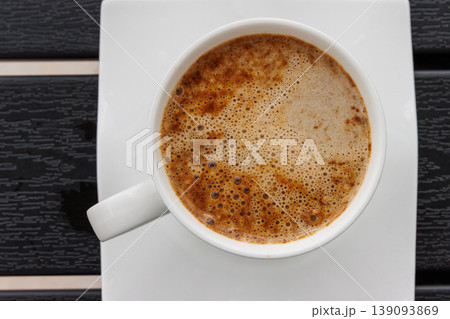 Close up top view of coffee cup on a square white saucer, placed on a dark slatted table. Selective focus. Copy space 139093869