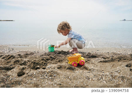 Little Boy Playing in Sand on Quiet Beach With Toy Truck and Bucket at Seaside Little Boy Playing in Sand on Quiet Beach With Toy Truck and Bucket at Seaside 139094758