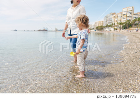 Mother and Young Child Walking on Sunny Beach Shore With City Skyline 139094778