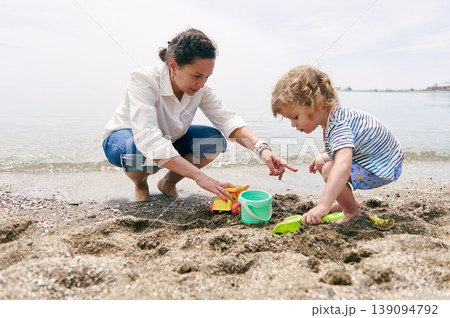 Mother and Toddler Playing on Sandy Beach With Bucket and Shovel on a Calm Summer Day 139094792