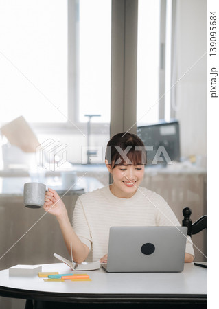 Happy asian woman working from home using technology for studying online business while drinking coffee holding mug smiling feeling relaxed and joyful looking at laptop screen sitting by window Happy asian woman working from home using technology for studying online business while drinking coffee holding mug smiling feeling relaxed and joyful looking at laptop screen sitting by window 139095684