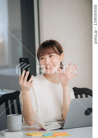 Cheerful smiling asian woman waving at smartphone camera during video call while happily working and studying online using modern technology for daily business routine from cozy home desk setup Cheerful smiling asian woman waving at smartphone camera during video call while happily working and studying online using modern technology for daily business routine from cozy home desk setup 139095688