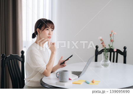 Thoughtful asian woman working home using modern technology studying online business strategy looking away contemplating future career success sitting at table with laptop and smartphone Thoughtful asian woman working home using modern technology studying online business strategy looking away contemplating future career success sitting at table with laptop and smartphone 139095690