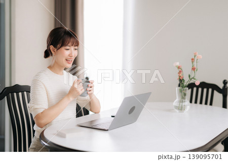 Happy asian woman drinking coffee from mug while working and studying online business at home using modern laptop technology feeling joyful smiling brightly sitting at white wooden table Happy asian woman drinking coffee from mug while working and studying online business at home using modern laptop technology feeling joyful smiling brightly sitting at white wooden table 139095701