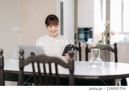 Happy young asian woman working from home studying online business technology sitting at dining table smiling looking at modern mobile device screen enjoying remote lifestyle Happy young asian woman working from home studying online business technology sitting at dining table smiling looking at modern mobile device screen enjoying remote lifestyle 139095704