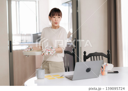 Focused asian woman at home working with technology while studying an online business course holding baking tray looking attentive at laptop screen standing near dining table Focused asian woman at home working with technology while studying an online business course holding baking tray looking attentive at laptop screen standing near dining table 139095724