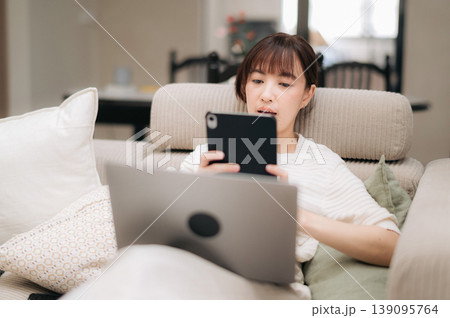 Focused asian woman comfortably working from home using modern technology while seriously studying online business concepts on laptop and mobile phone resting on soft living room sofa Focused asian woman comfortably working from home using modern technology while seriously studying online business concepts on laptop and mobile phone resting on soft living room sofa 139095764