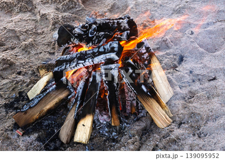 Burning campfire on a sandy beach. Bright orange flames and charred wood logs. Outdoor camping and survival concept Burning campfire on a sandy beach. Bright orange flames and charred wood logs. Outdoor camping and survival concept 139095952