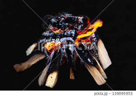 Burning campfire with bright yellow flames and glowing red embers. Charred wood logs isolated on a dark black background. Burning campfire with bright yellow flames and glowing red embers. Charred wood logs isolated on a dark black background. 139095953