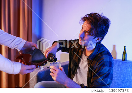 Depressed drunk man pouring alcohol into a glass on the sofa while a person holds an empty wallet towards him. Concept of poverty, unemployment, alcoholism, and financial crisis. Depressed drunk man pouring alcohol into a glass on the sofa while a person holds an empty wallet towards him. Concept of poverty, unemployment, alcoholism, and financial crisis. 139097578