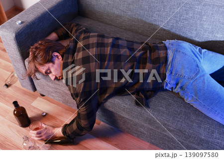 Unconscious drunk man sleeping on a sofa at home, holding a glass. Empty alcohol bottles on the floor. Concept of addiction, unemployment, divorce, or severe depression. Unconscious drunk man sleeping on a sofa at home, holding a glass. Empty alcohol bottles on the floor. Concept of addiction, unemployment, divorce, or severe depression. 139097580