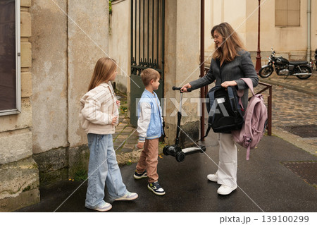 Mother meeting children at courtyard gate with scooter after school. Back to school Mother meeting children at courtyard gate with scooter after school. Back to school 139100299