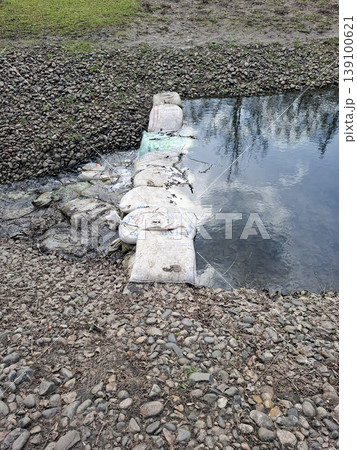 A small dam is built across a narrow stream using white sandbags. The setting is a rural or park area with visible stones and grass nearby. Water flows over the dam. A small dam is built across a narrow stream using white sandbags. The setting is a rural or park area with visible stones and grass nearby. Water flows over the dam. 139100621