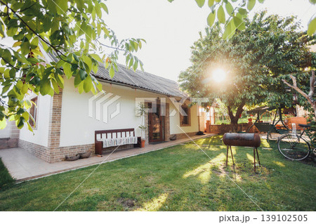 Sunlit Backyard With Bicycle And Barbecue, Warm Golden Light Filtering Through Trees Over Grass And Bench Mood Evokes Family Leisure, Evening Picnic, Relaxed Summer Lifestyle Scenes. 139102505