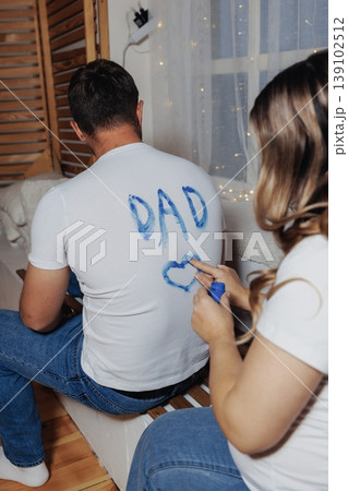 Woman Painting DAD On Shirt Behind Seated Partner, Brush Forming Playful Blue Letters And Heart, Shuttered Wooden Backdrop With String Lights, Cushions And Cozy Textures, Close Focus 139102512