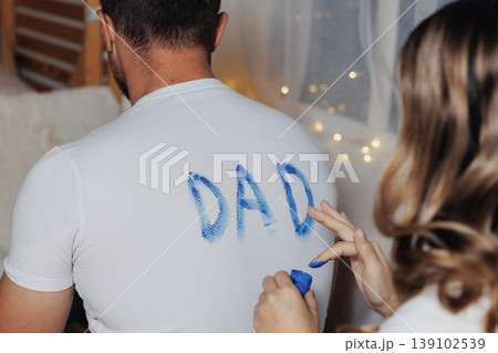 Man In White Shirt Painted DAD, Woman Applying Blue Paint Heart On Lower Back, Cozy Room With Fairy Lights, Wooden Floor, Casual Jeans, Playful Intimate Moment, DIY Gift Preparation 139102539