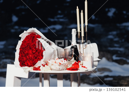 Red Rose Bouquet On White Table, Champagne Bottle, Scattered Petals And Folded Note Create Festive Romantic Scene With Elegant Candelabra, Polished Glassware And Crisp Snowy Backdrop 139102541